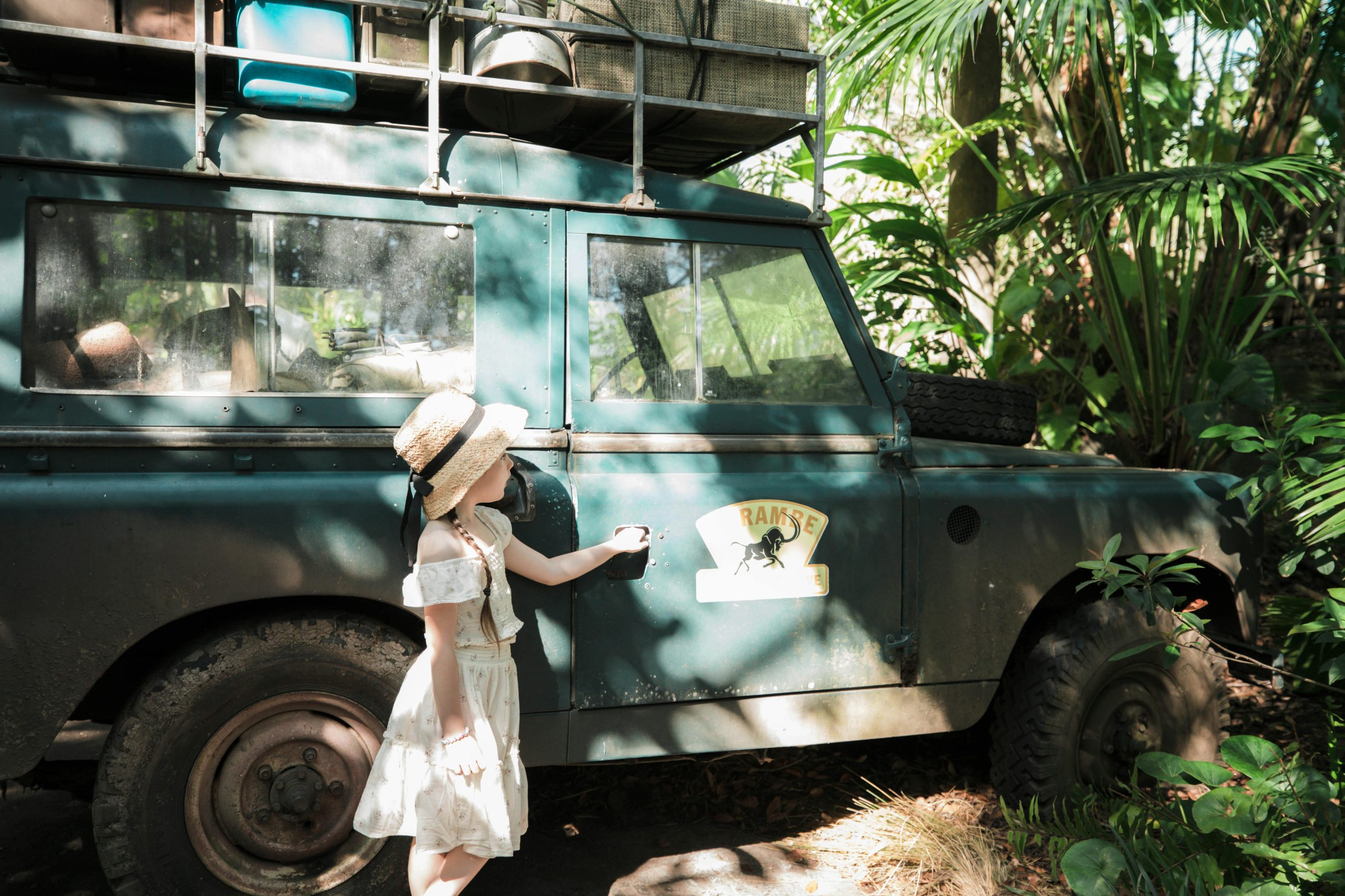 A young girl in a hat stands next to a vintage safari jeep in a lush jungle setting.